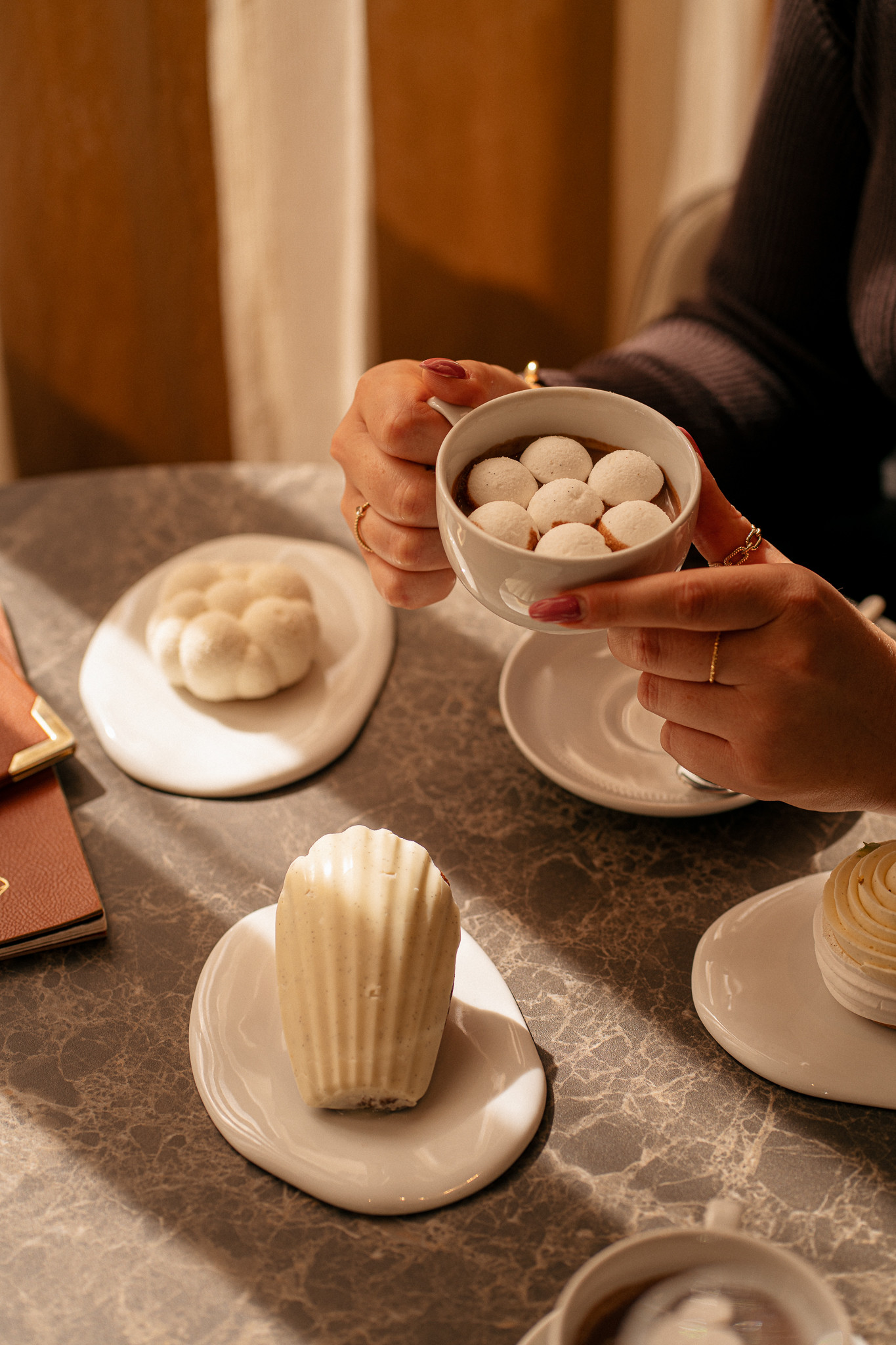 Personne tenant une tasse contenant des meringues blanches, avec des desserts élégants sur une table en marbre.