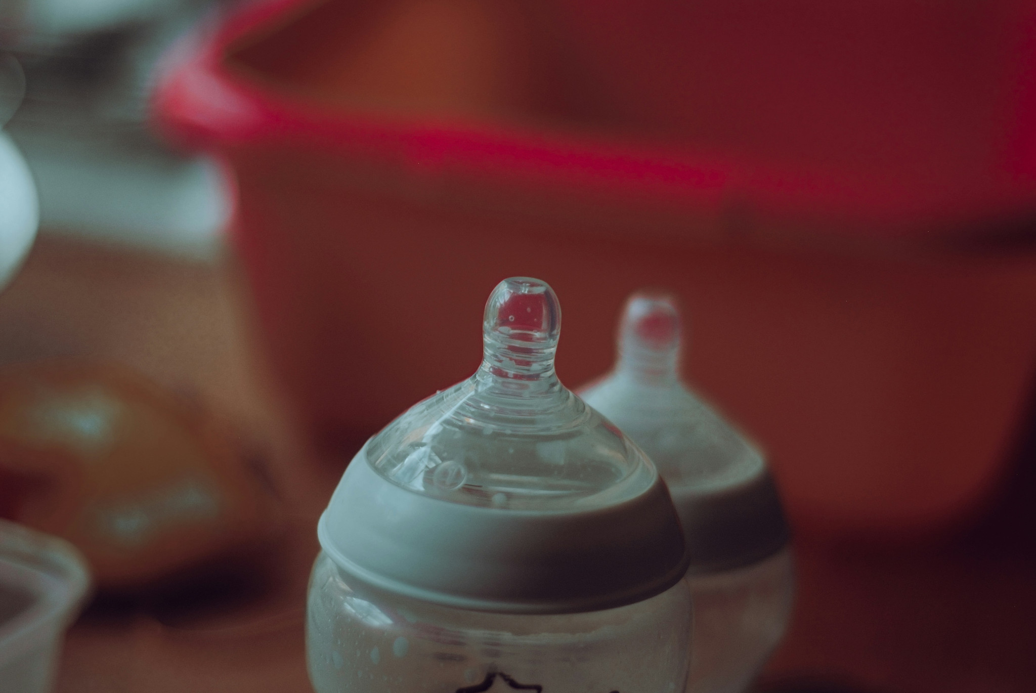 Deux biberons en plastique transparent avec tétines en silicone, posés sur une surface, avec un récipient rouge en arrière-plan.