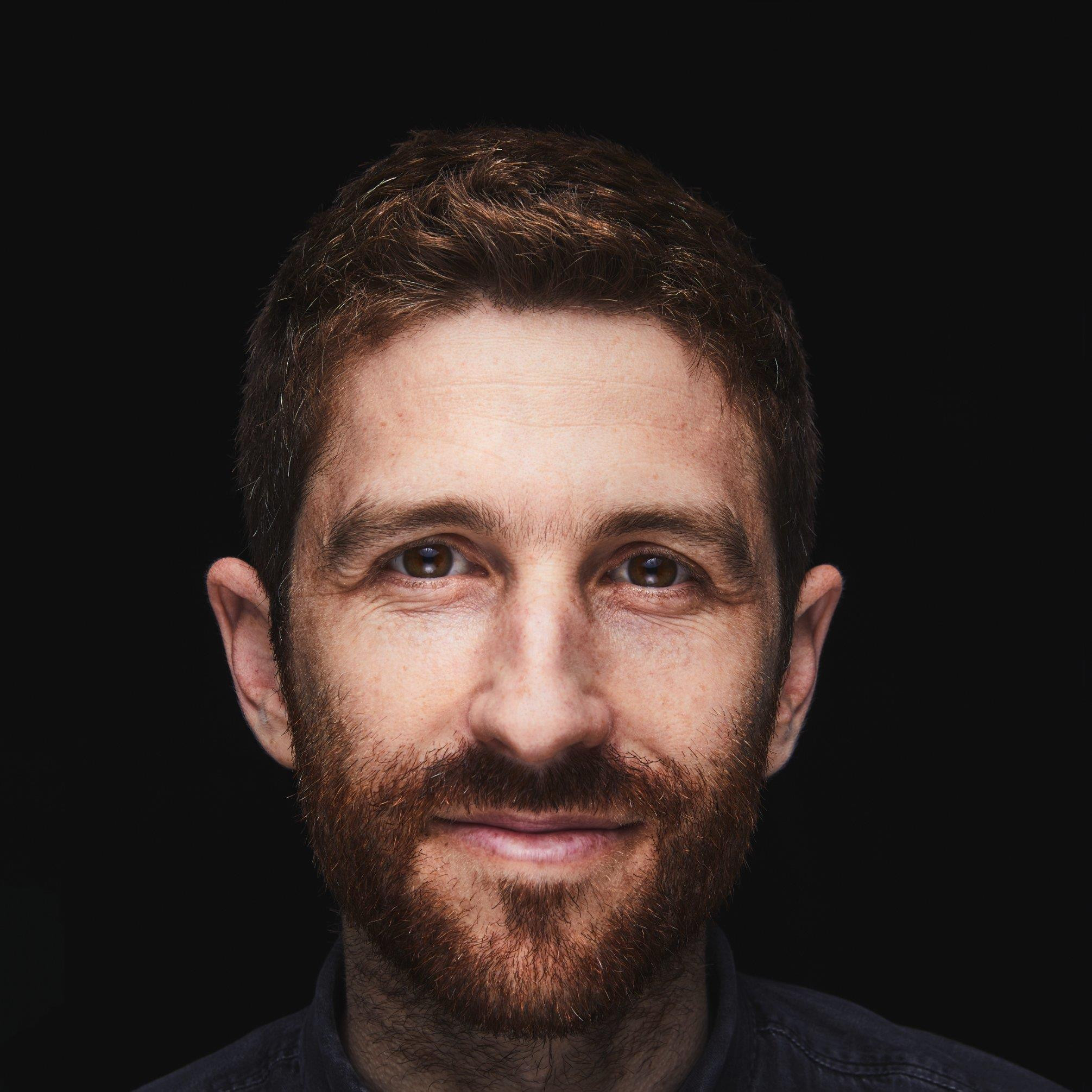 Portrait d’un homme aux cheveux et à la barbe châtain roux, souriant légèrement, sur fond noir.