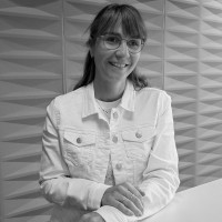 Femme souriante portant une veste en jean blanche contre un mur texturé en noir et blanc.
