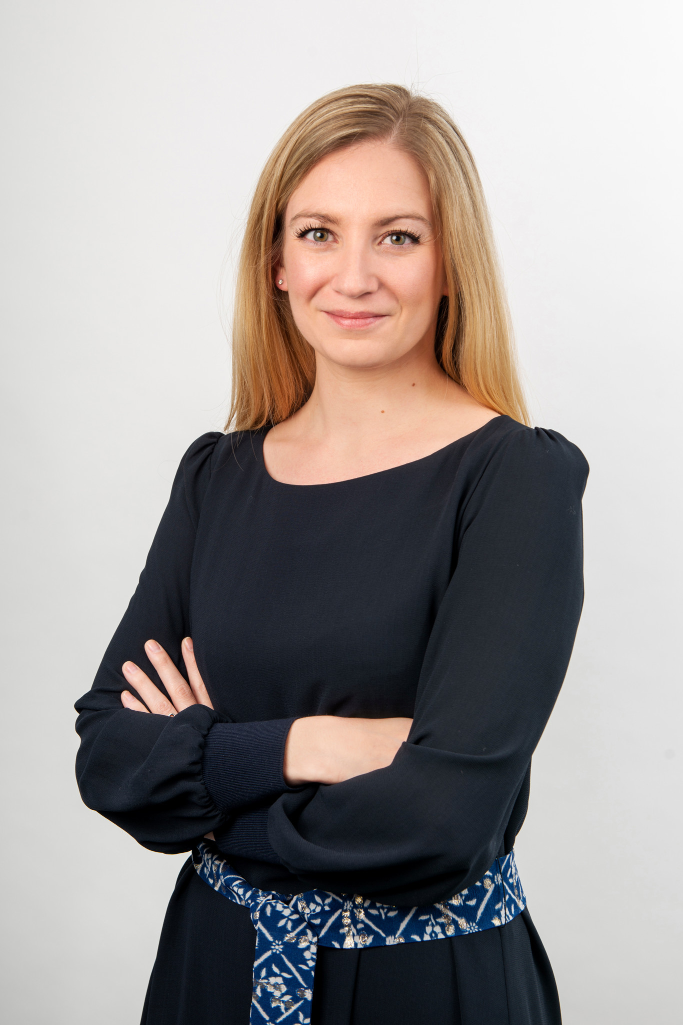 Femme blonde souriante en haut noir, bras croisés, avec une ceinture à motifs bleus, sur fond blanc neutre.