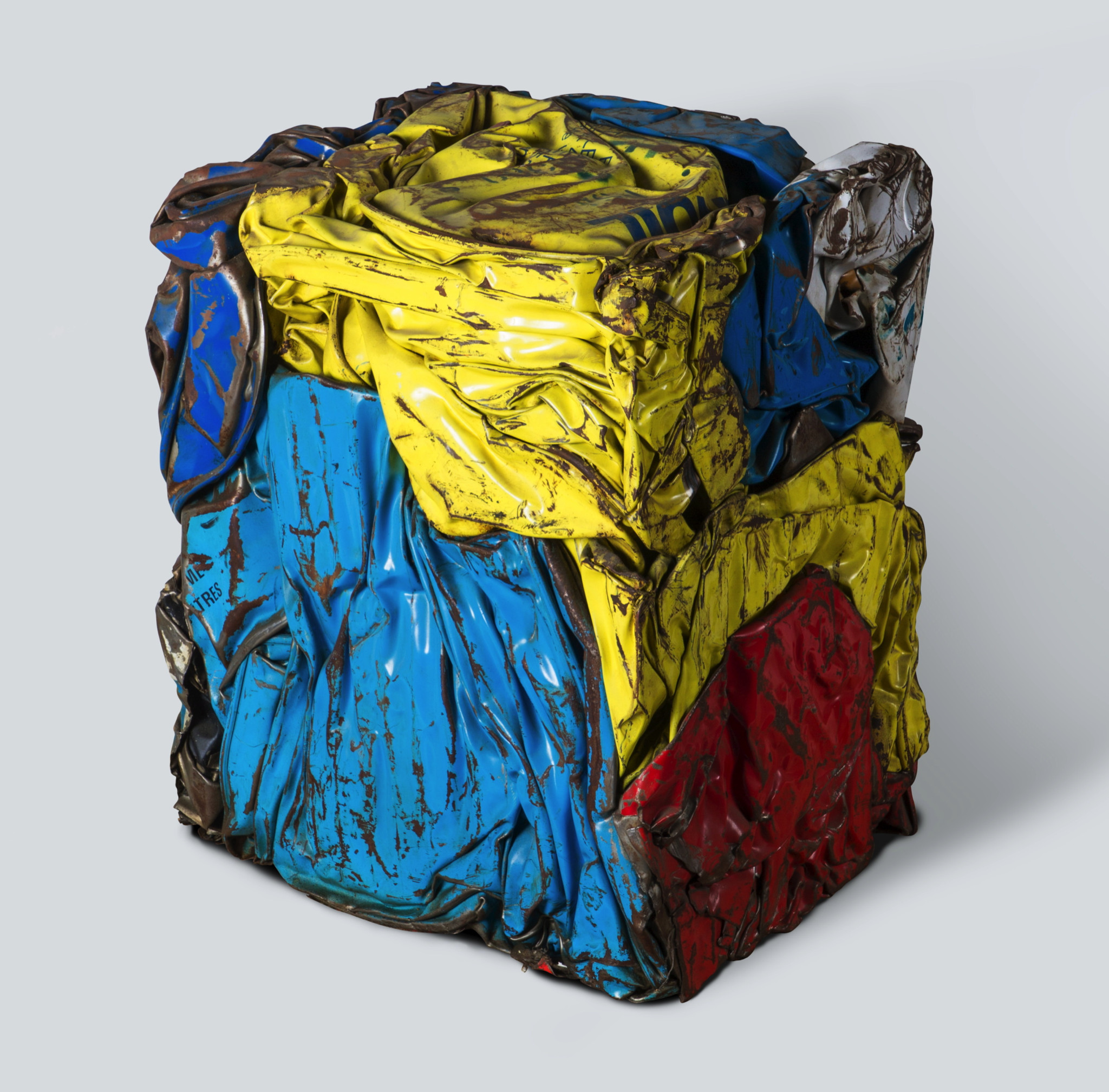 Sculpture cubique en métal compressé aux couleurs vives: bleu, jaune et rouge, aux textures froissées, sur fond gris neutre.