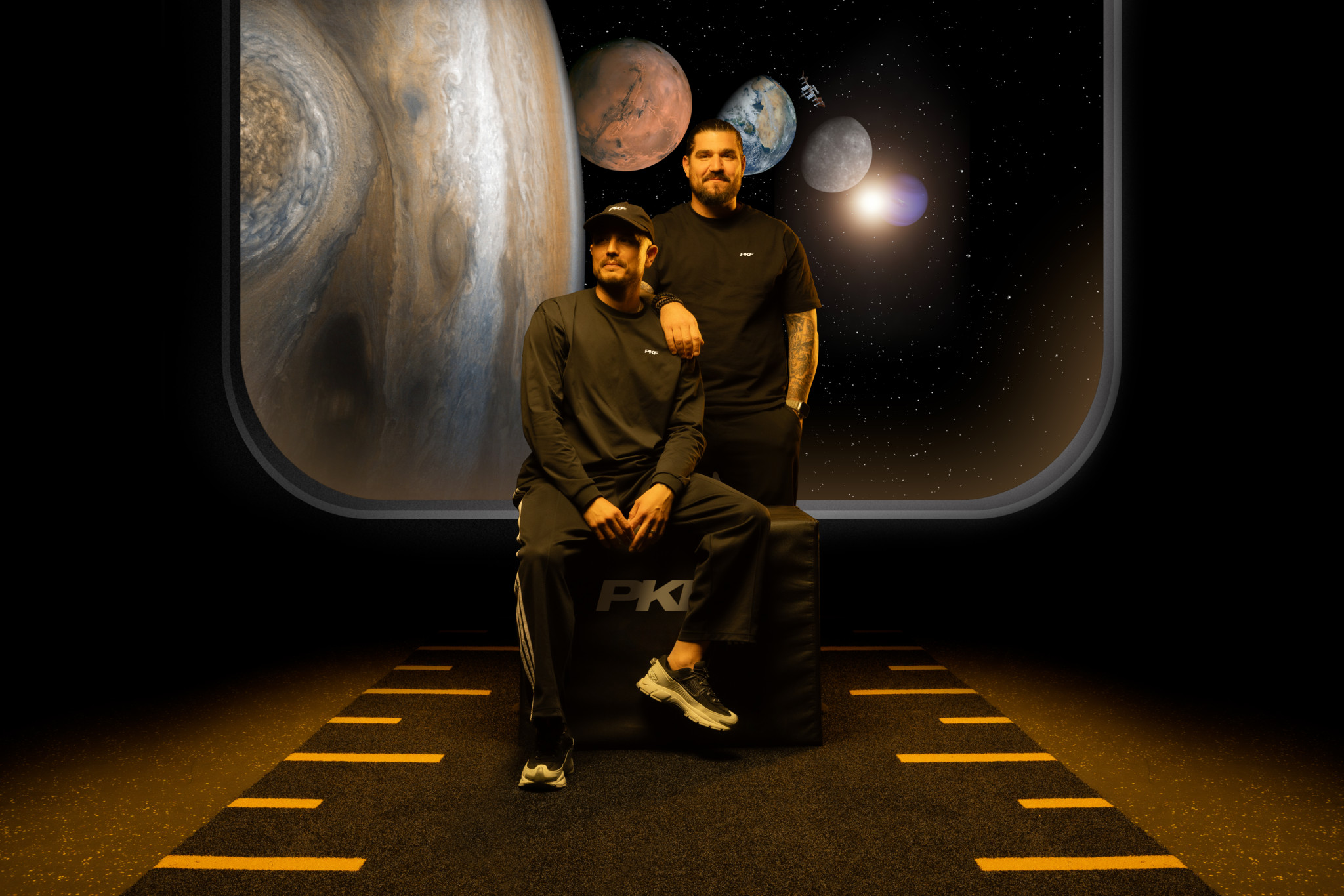 Deux hommes en tenue sombre posent devant un décor spatial avec planètes, dont Jupiter, visible par un hublot stylisé.