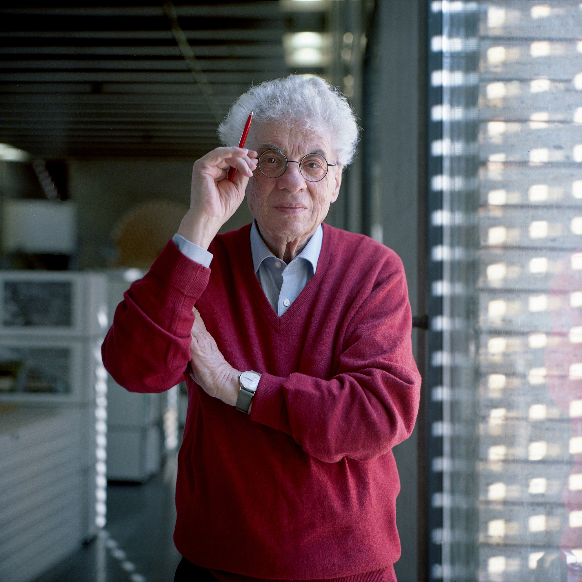 Un homme âgé, portant un pull rouge et des lunettes, tient un crayon rouge et se tient dans un bureau moderne.