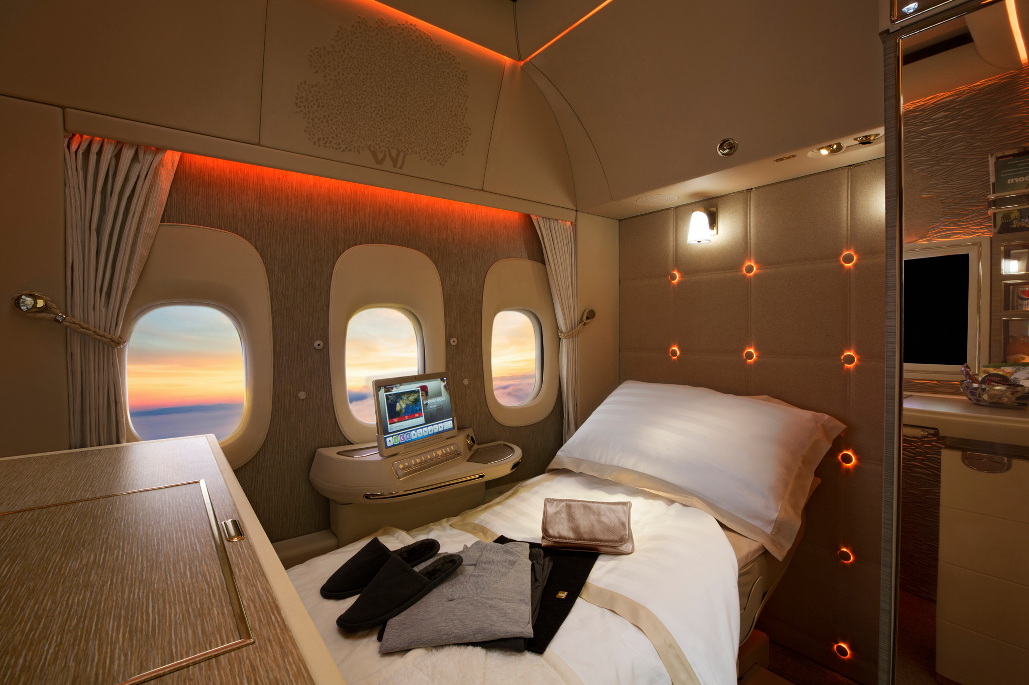 Intérieur luxueux d’une cabine d’avion avec un lit, un écran et des fenêtres offrant une vue sur le coucher de soleil.
