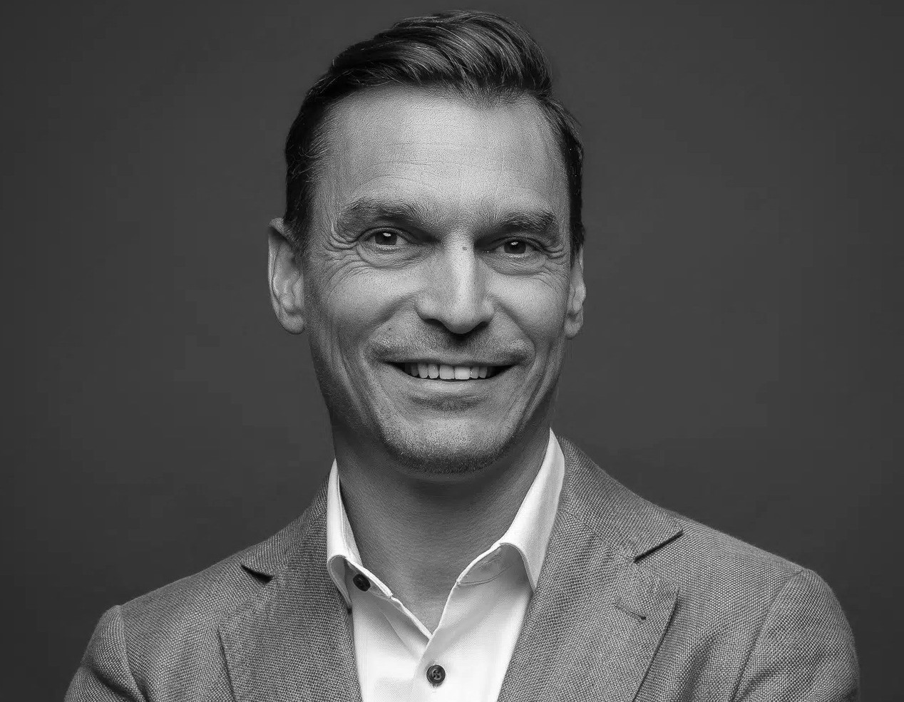 Portrait en noir et blanc d’un homme souriant en veste et chemise blanche sur fond gris.