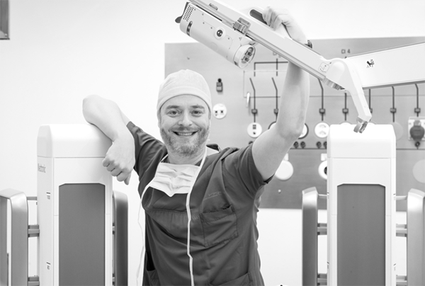 Un chirurgien souriant en tenue médicale pose avec les bras d’un robot chirurgical dans une salle d’opération.