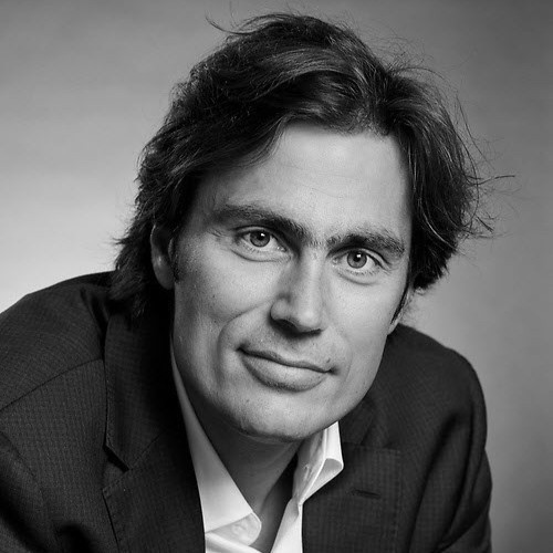 Portrait en noir et blanc d’un homme aux cheveux bruns ondulés, vêtu d’un veston sombre et d’une chemise blanche, souriant légèrement.
