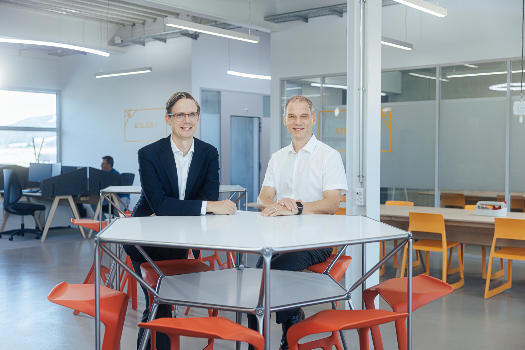 Deux hommes souriants assis à une table blanche dans un espace de bureau moderne avec des chaises rouges et oranges.