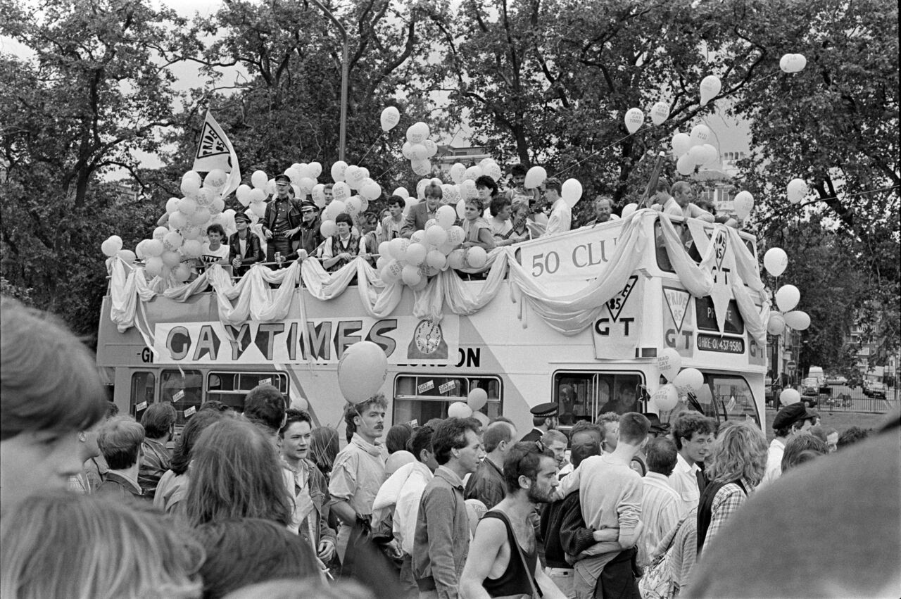 Un bus à impériale décoré de ballons avec l’inscription ’Gay Times’ lors d’une parade festive, entouré d’une foule nombreuse.