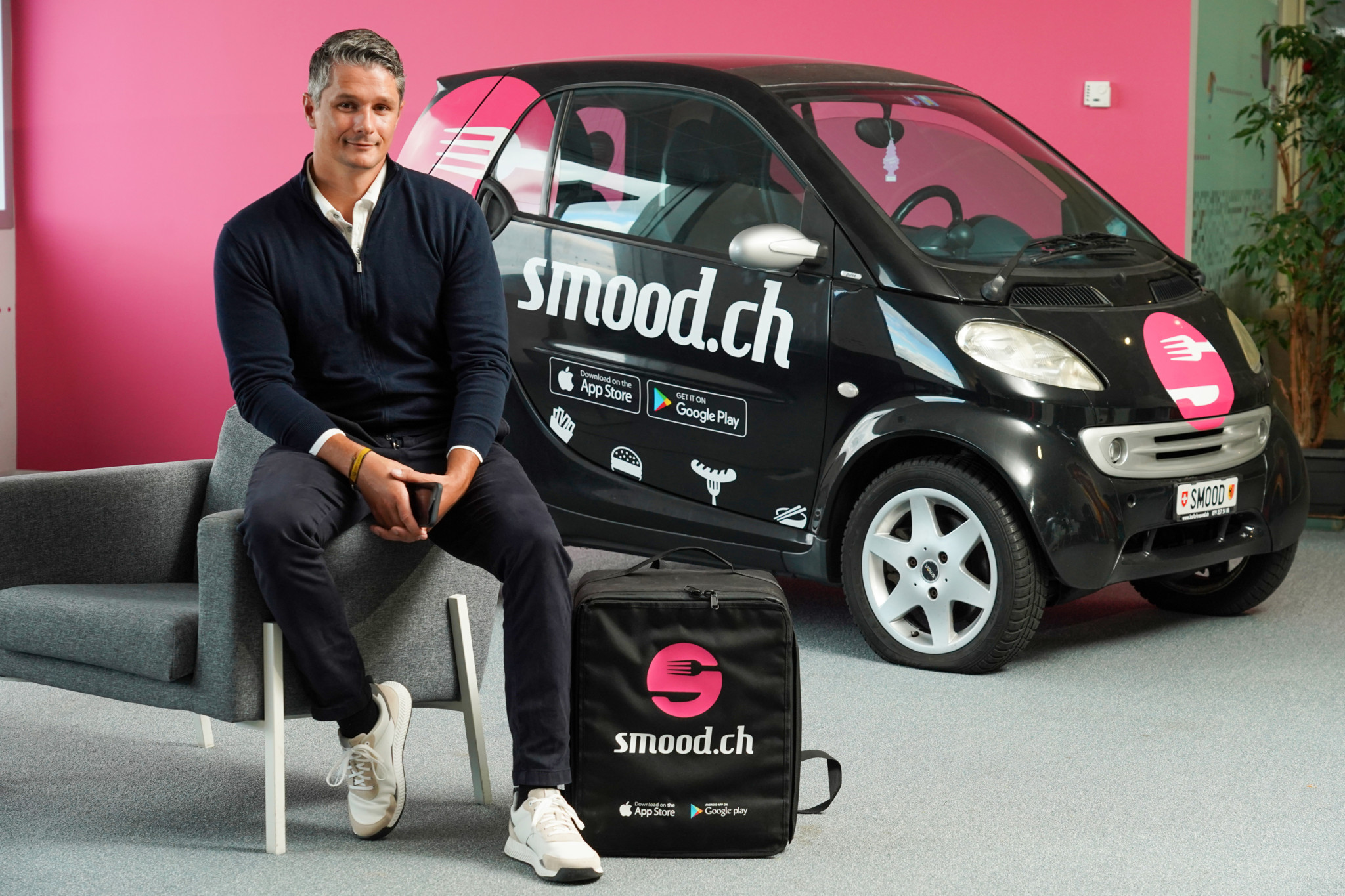 Marc Aeschlimann, fondateur de Smood, assis devant une Smart noire frappée du logo smood.ch, avec un sac de livraison à ses pieds.