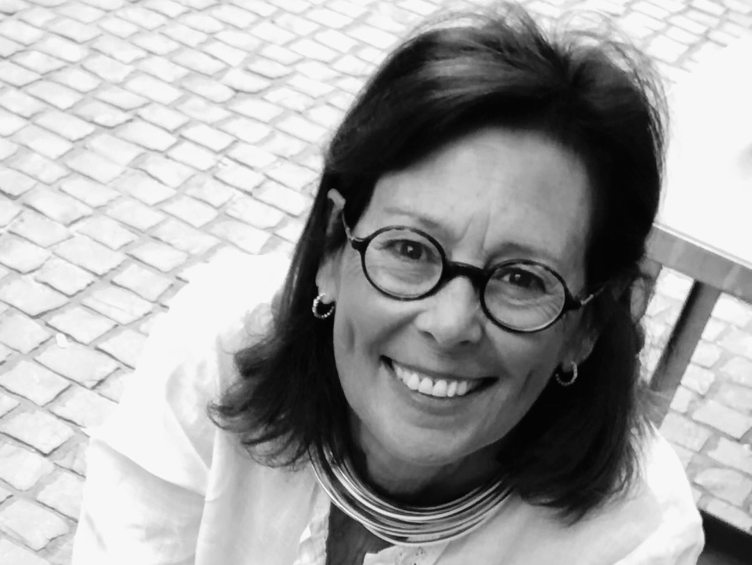 Femme souriante aux cheveux bruns et lunettes rondes, photographiée en noir et blanc sur fond de pavés.
