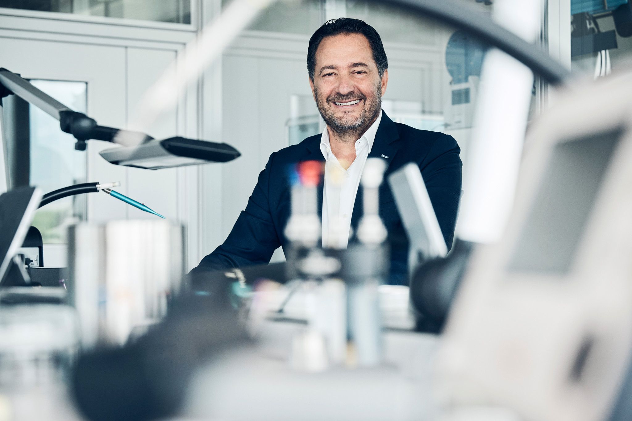 Homme souriant en veste sombre assis à un établi d’horlogerie, entouré d’outils de précision et d’équipements de laboratoire.