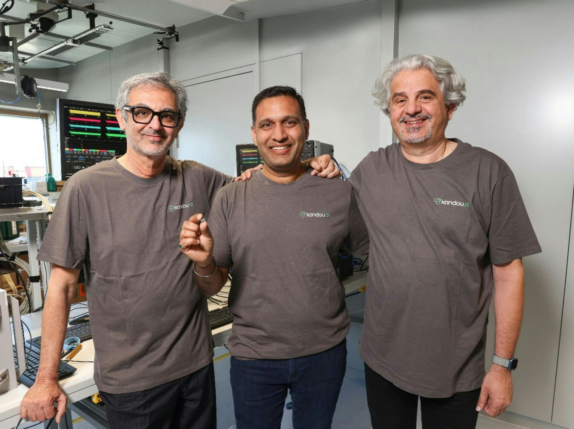 Trois hommes souriants portant des t-shirts Sandou posent ensemble dans un laboratoire, l’un tenant un petit composant électronique.