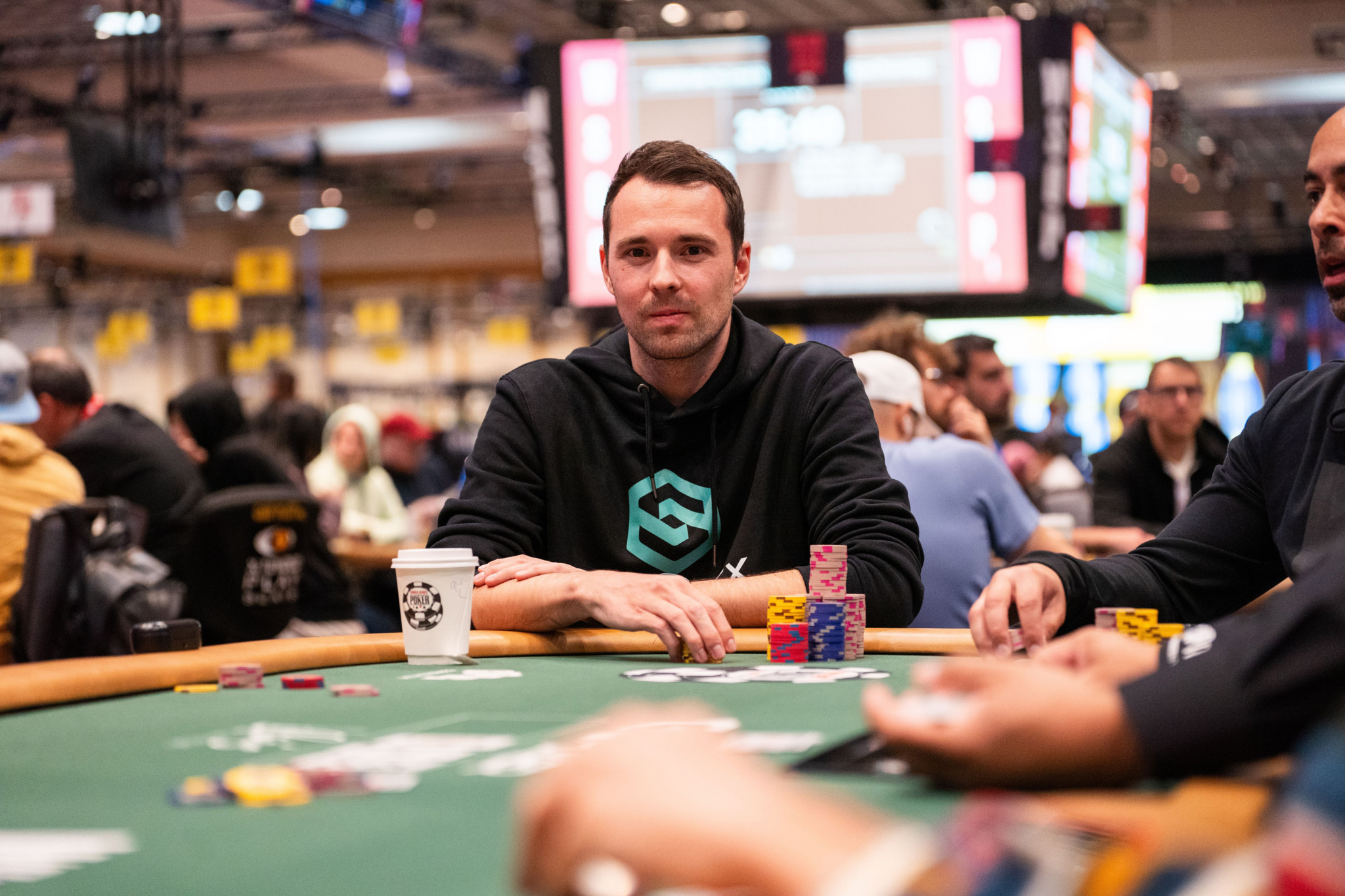 Un homme joue au poker dans un casino, entouré de plusieurs autres joueurs et de jetons de poker colorés sur la table.