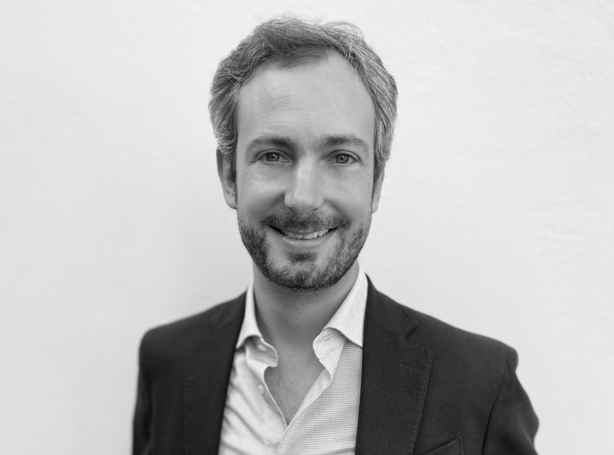 Portrait en noir et blanc d’un homme souriant en veste sombre et chemise claire, sur fond blanc.