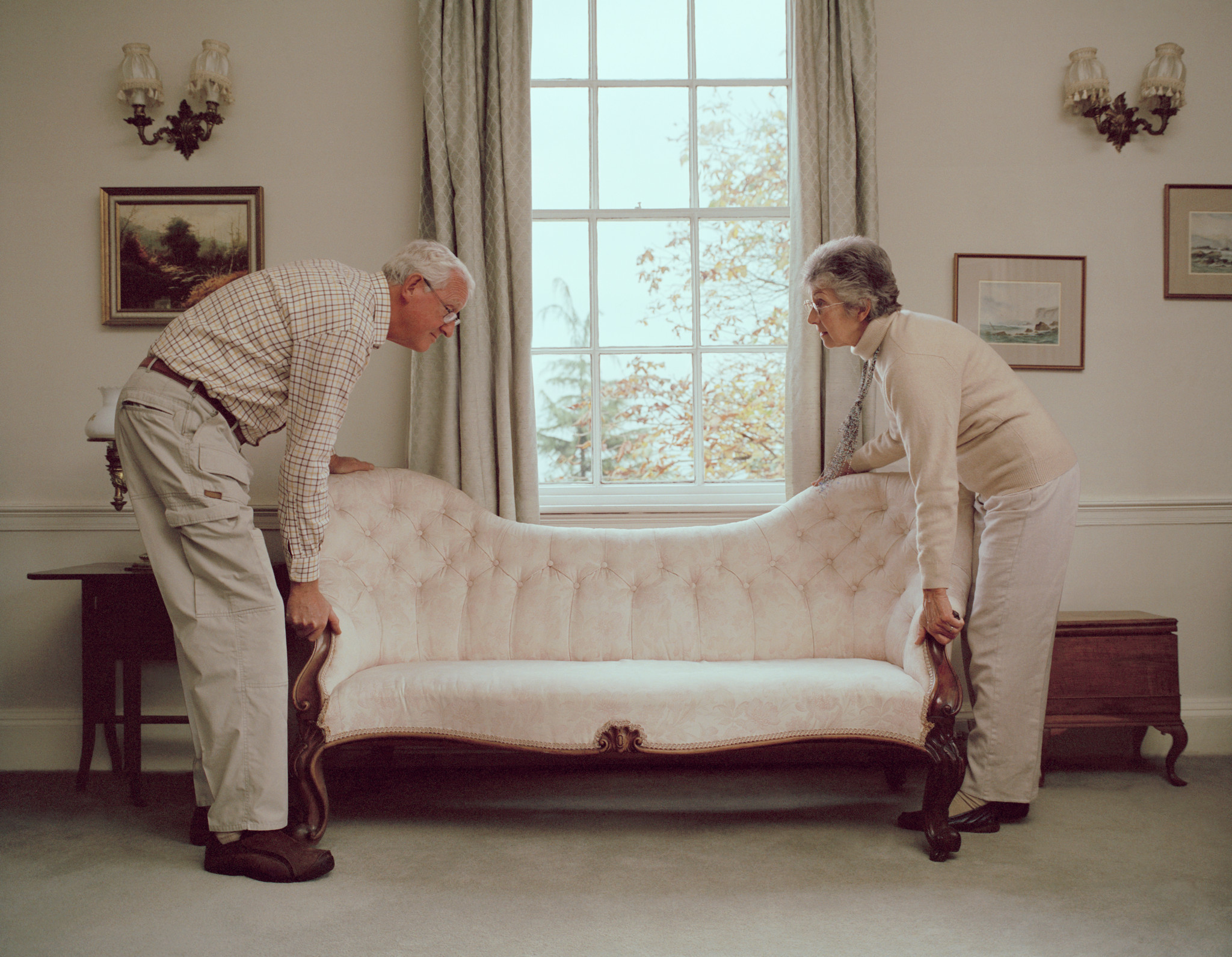 Un couple âgé déplace un canapé ancien dans une pièce lumineuse avec une grande fenêtre.