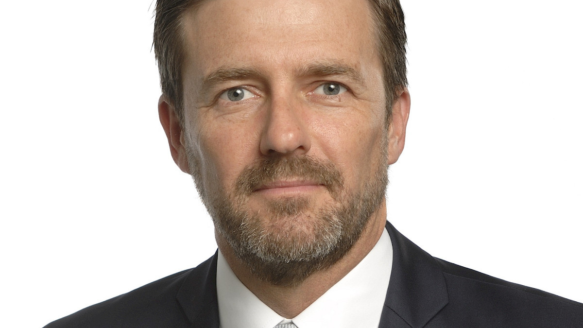 Portrait d’un homme en costume sombre et cravate, barbe courte, regard direct, sur fond blanc.