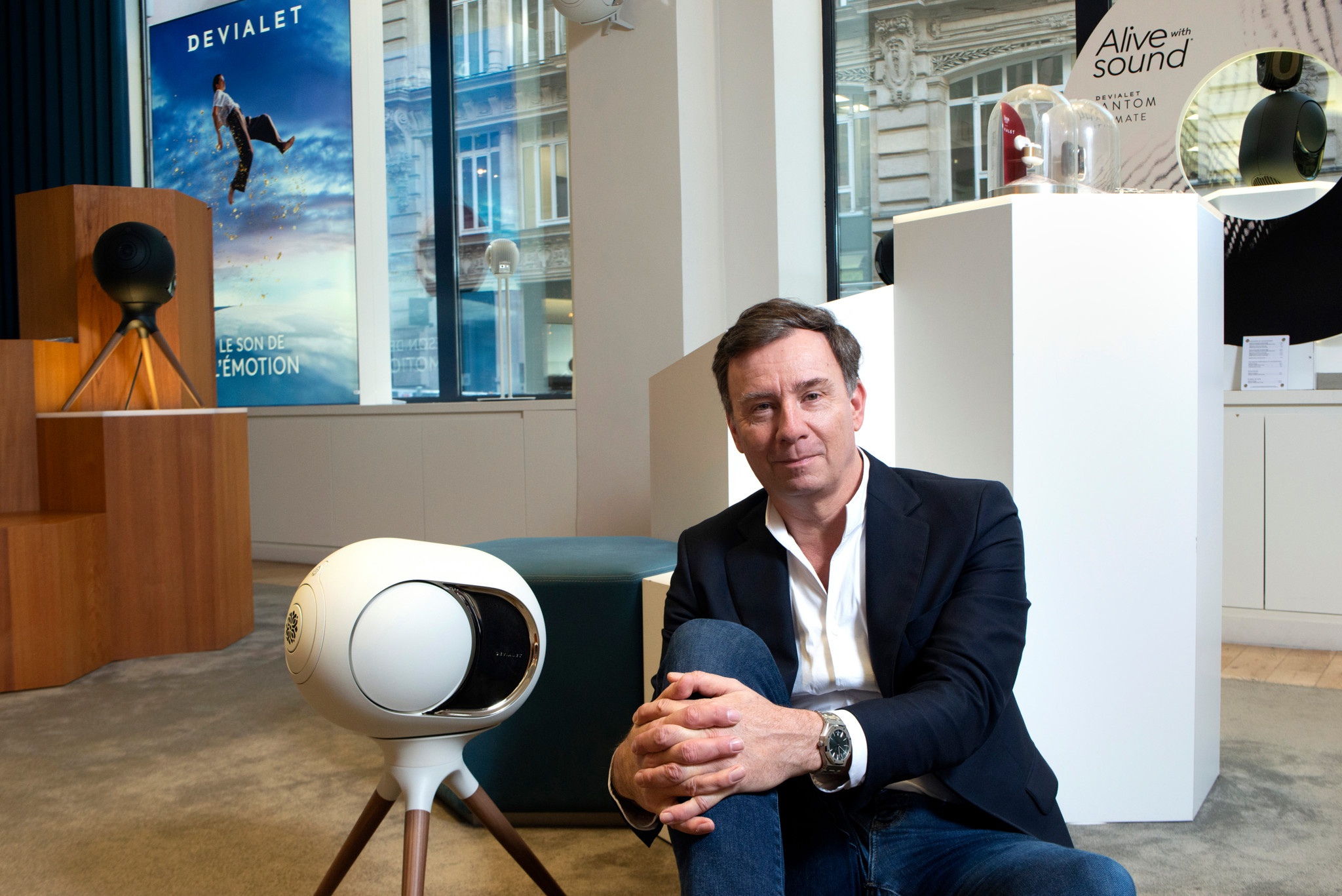 Jacques Demont, CEO de Devialet, assis dans un showroom avec des enceintes et affiches de la marque en arrière-plan.