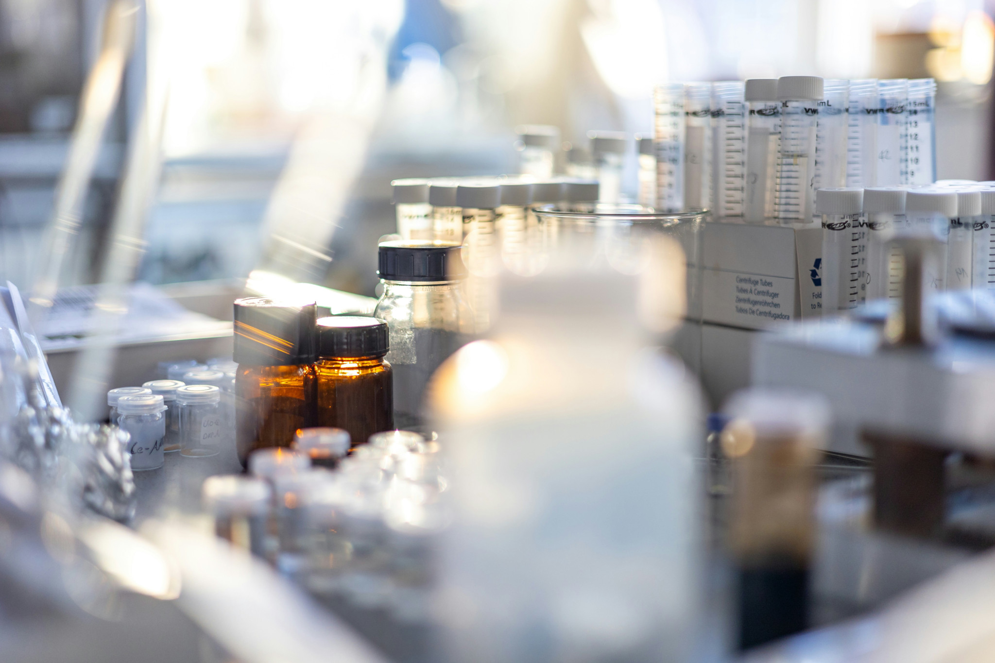 Flacons et tubes de laboratoire pharmaceutique alignés sur un plan de travail, dont un flacon ambré à bouchon noir au premier plan.