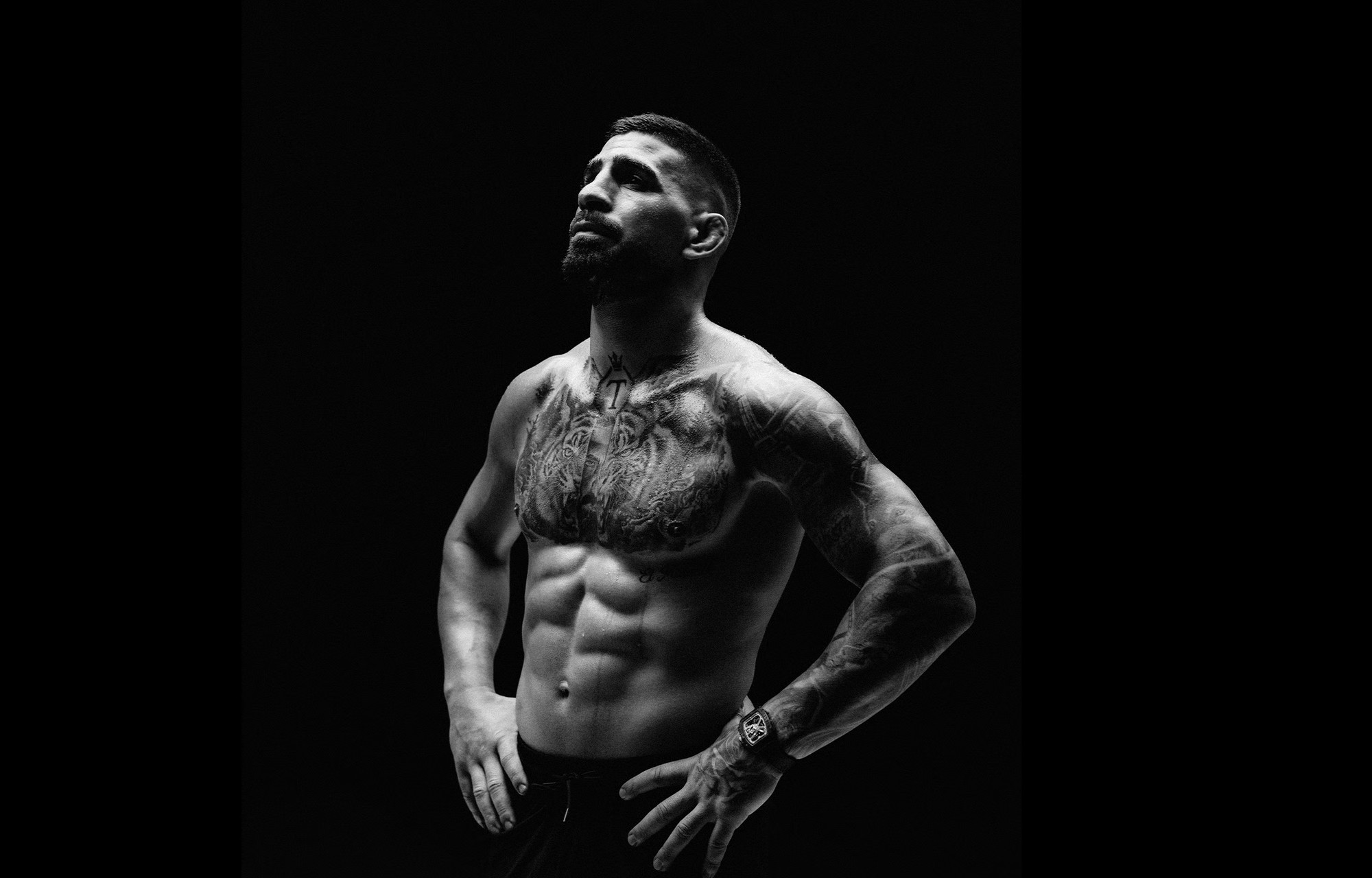 Homme musclé torse nu avec tatouages sur la poitrine, pose dramatique en noir et blanc sur fond sombre.