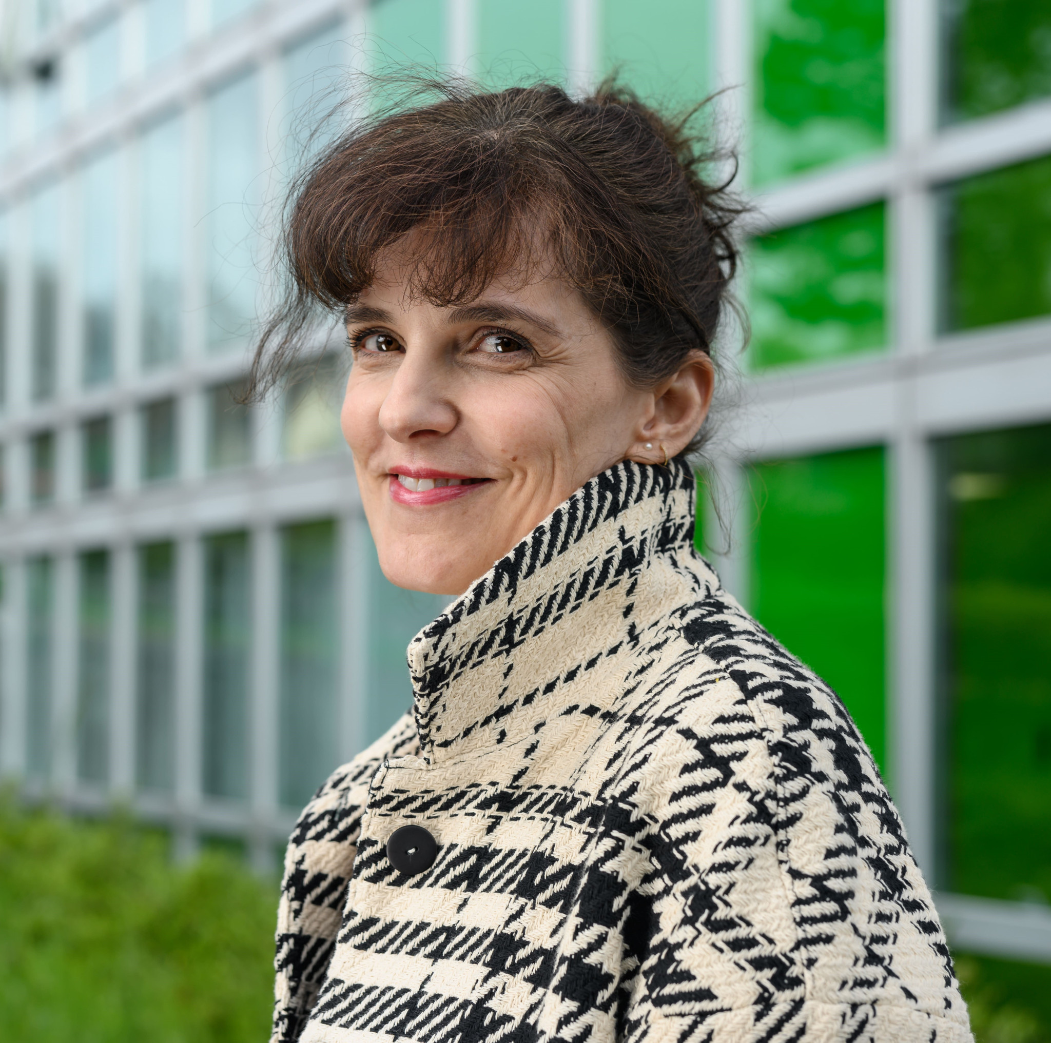 Femme souriante en manteau à motifs devant un bâtiment moderne avec des fenêtres et des reflets verts.