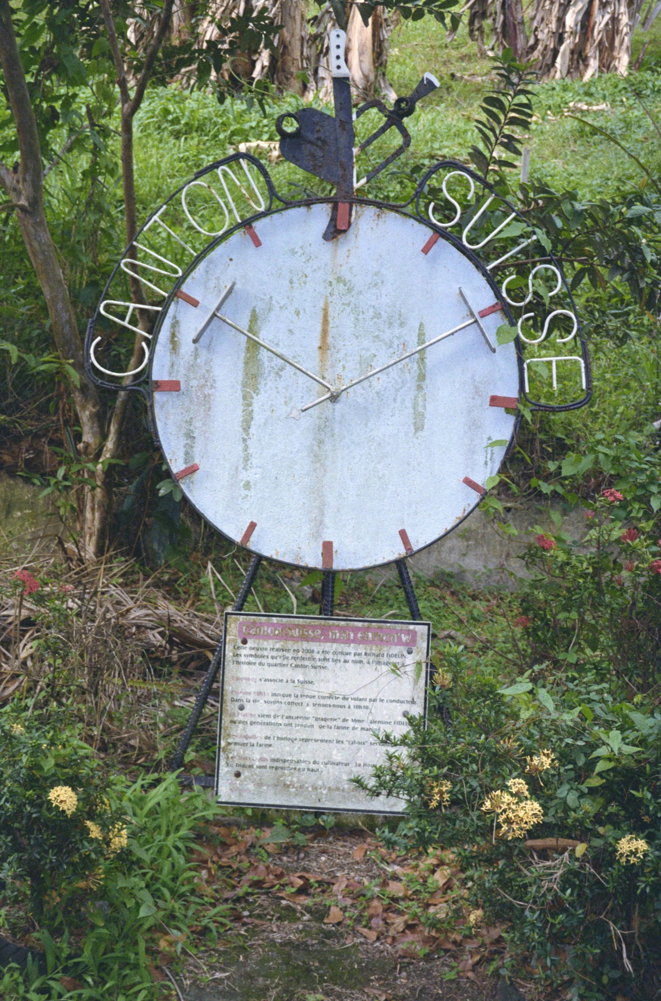 Grande horloge métallique vintage avec l’inscription ’Canton Suisse’, entourée de végétation tropicale et fleurs colorées, avec un panneau informatif.