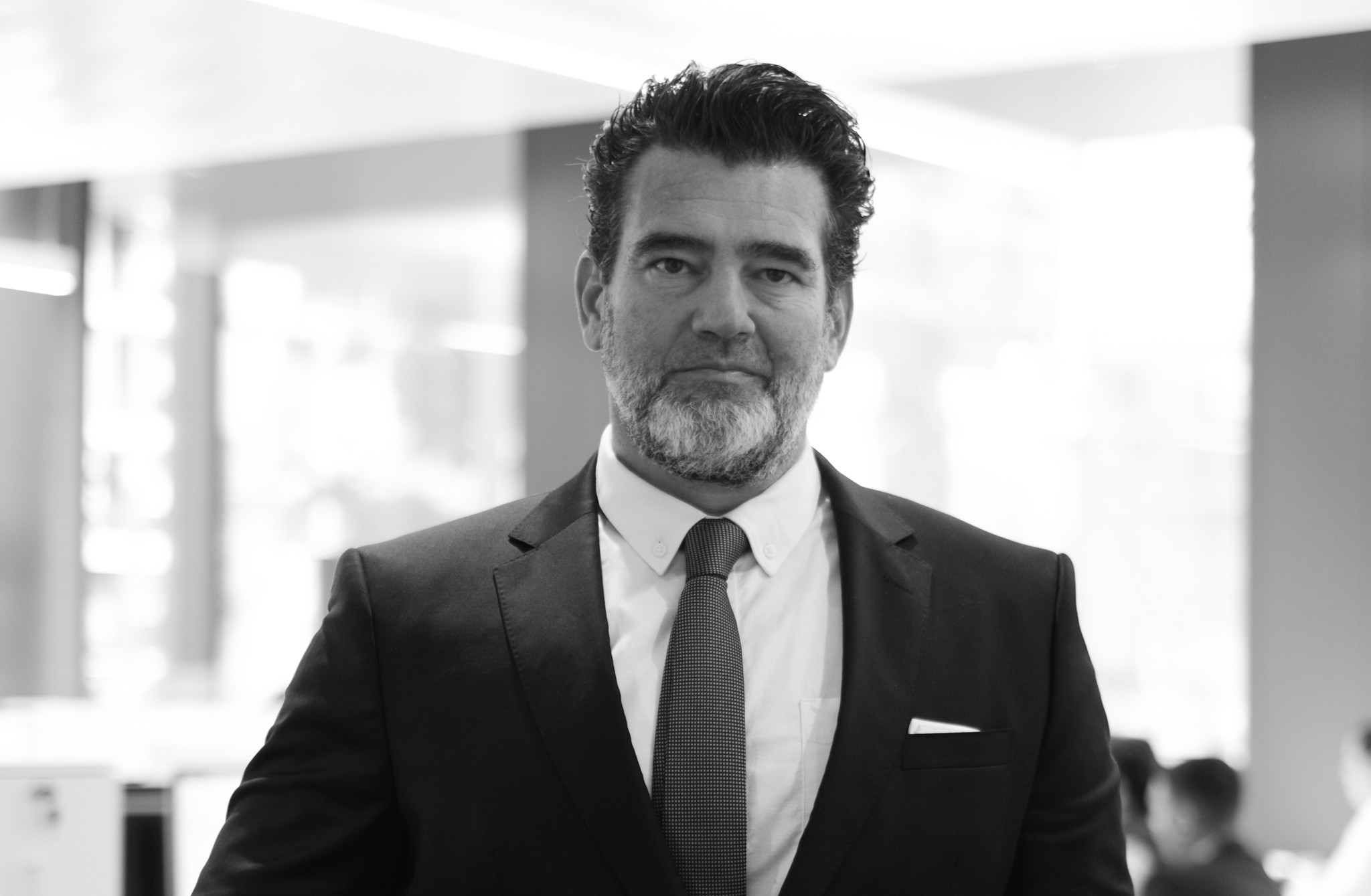 Homme en costume sombre et cravate, barbe courte, pose sérieuse, photo en noir et blanc dans un environnement professionnel.