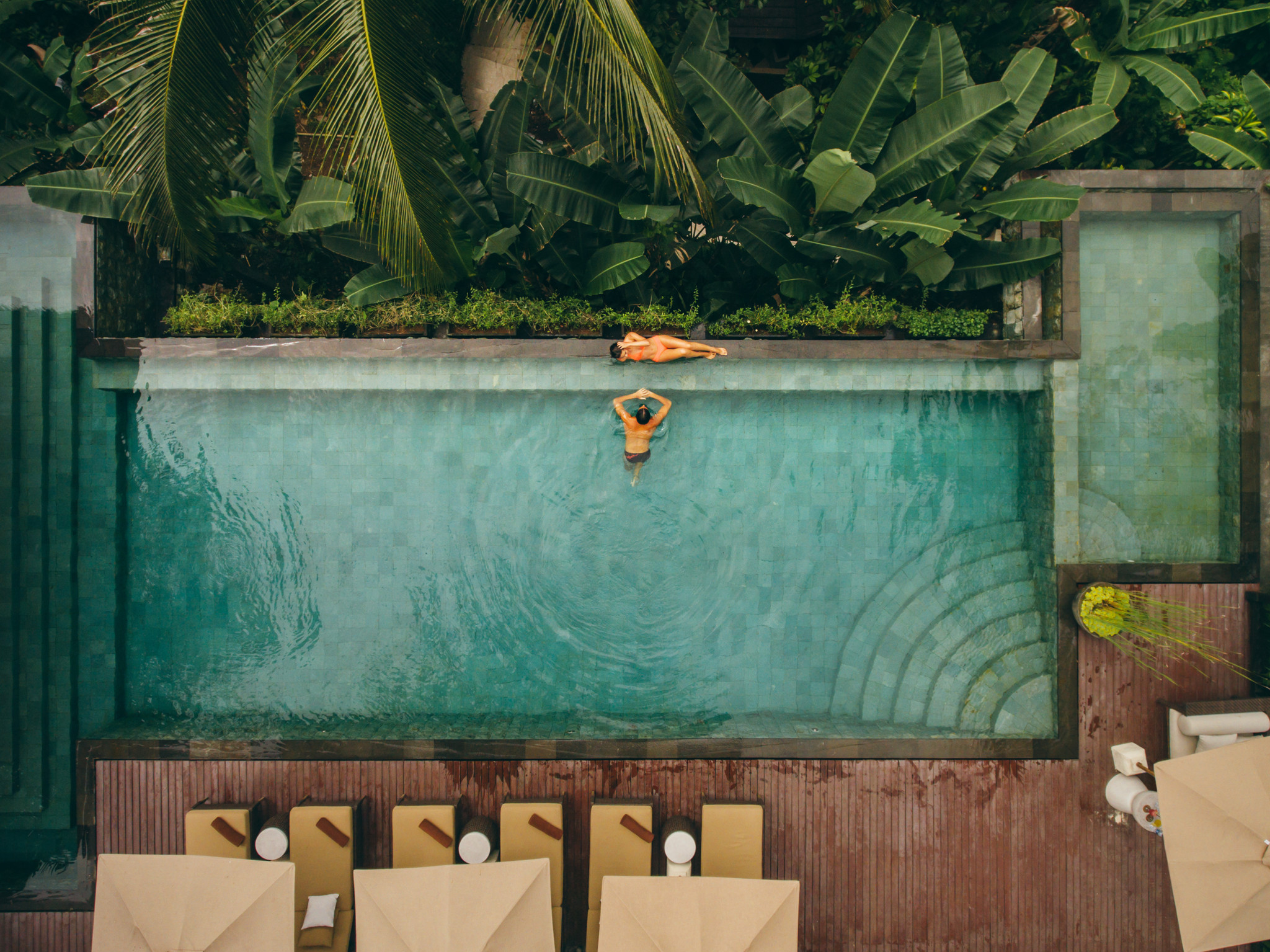 Vue aérienne d’un couple en vacances dans un resort de luxe: une femme se détend au bord de la piscine turquoise, un homme nage, entourés de végétation tropicale.
