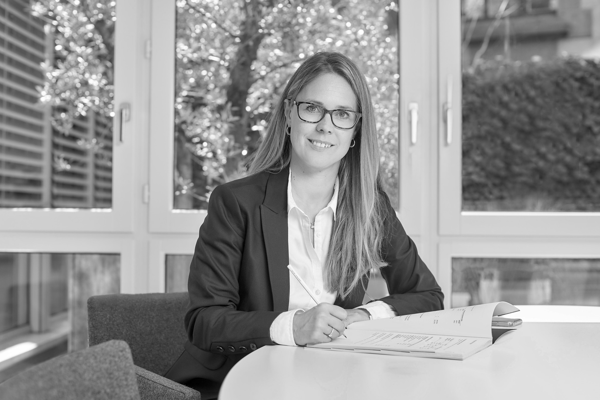 Femme en veste sombre et lunettes, assise à un bureau avec des documents, souriant devant une fenêtre avec arbres en arrière-plan.