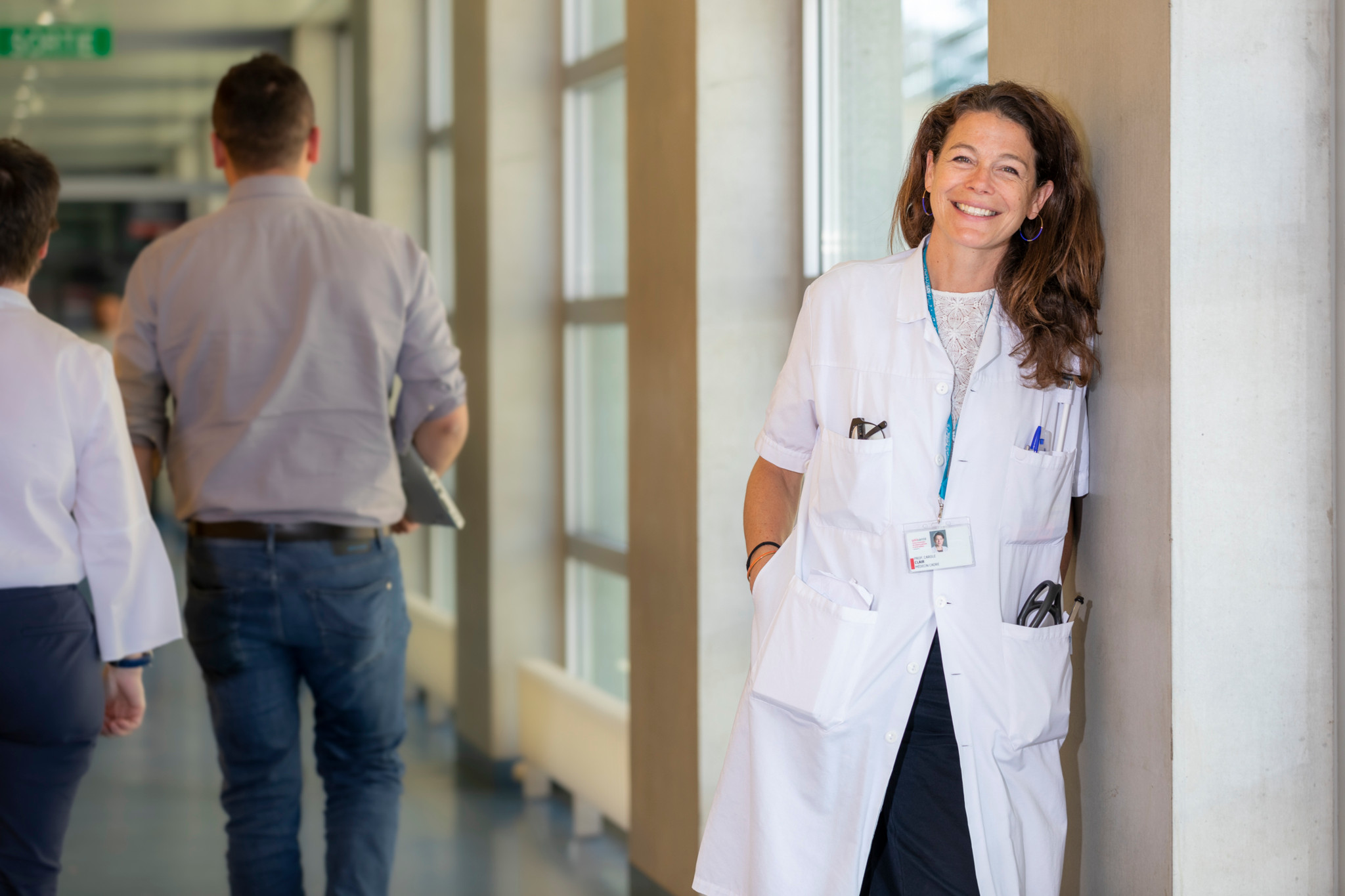 Médecin souriante en blouse blanche appuyée contre un mur dans un couloir d’hôpital lumineux.