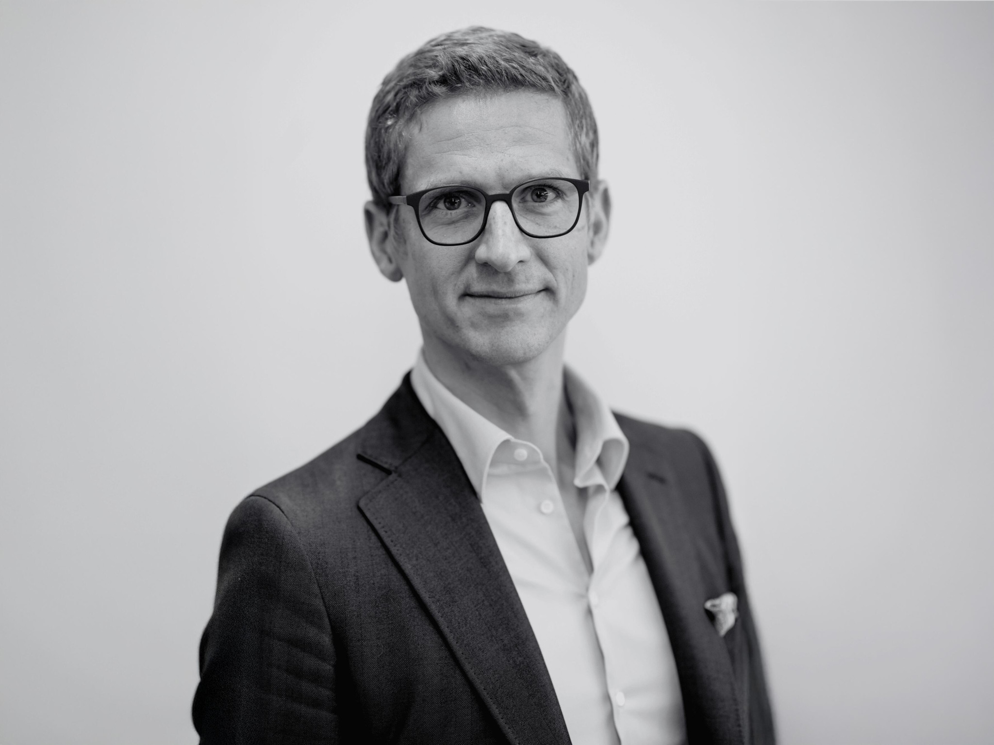 Portrait en noir et blanc d’un homme en veste sombre et chemise blanche, portant des lunettes, sur fond neutre.