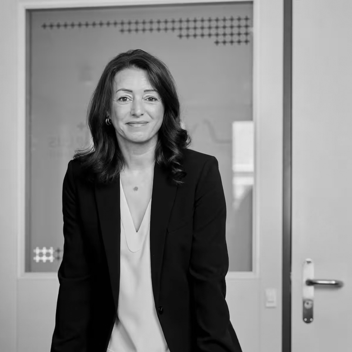 Femme en veste noire souriante devant une porte vitrée de bureau, portrait professionnel en noir et blanc