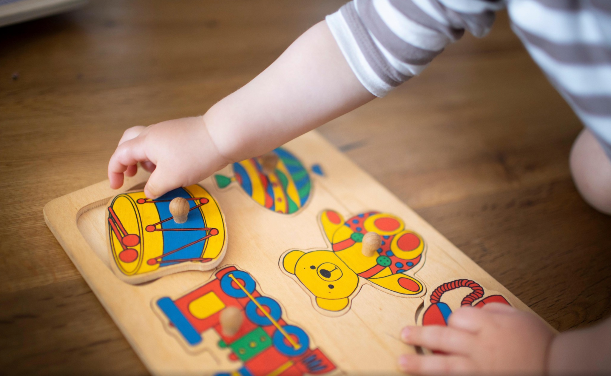 Jeune enfant jouant avec un puzzle en bois coloré représentant des jouets : tambour, ours en peluche et train.