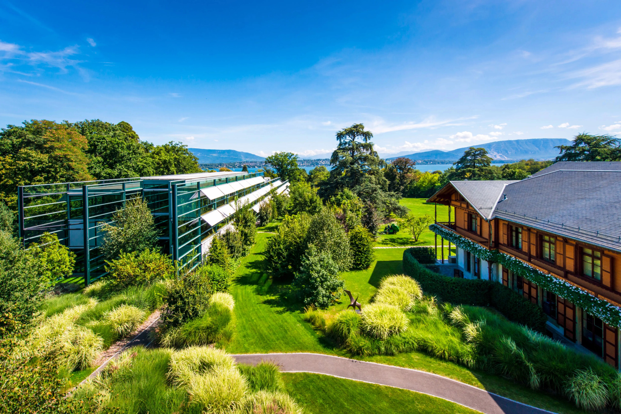 Vue aérienne d’un domaine élégant avec jardins verdoyants, bâtiment moderne vitré et villa en bois, face à un lac sous ciel bleu.
