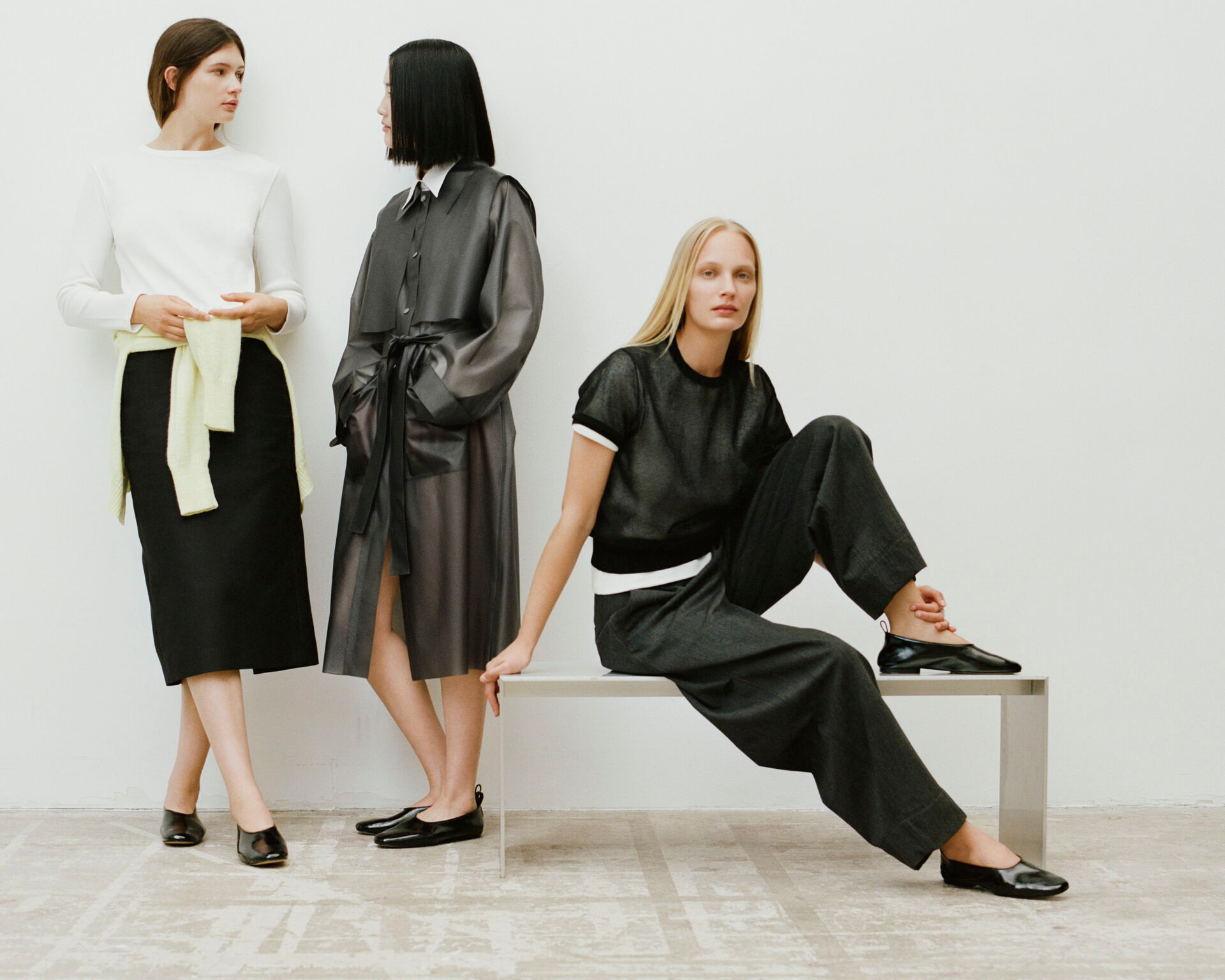 Trois mannequins en tenues minimalistes noir et blanc, dont une robe en cuir transparent et un ensemble pantalon, posant dans un cadre épuré.