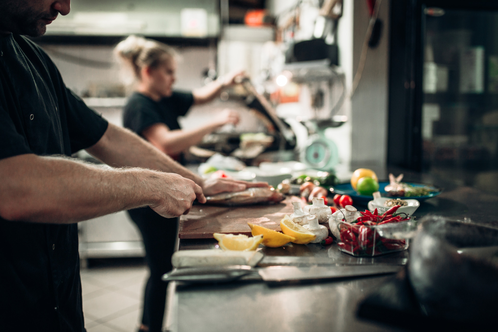 Deux cuisiniers préparent des spécialités de la mer dans une cuisine de restaurant, avec poissons, citrons et piments sur le plan de travail.