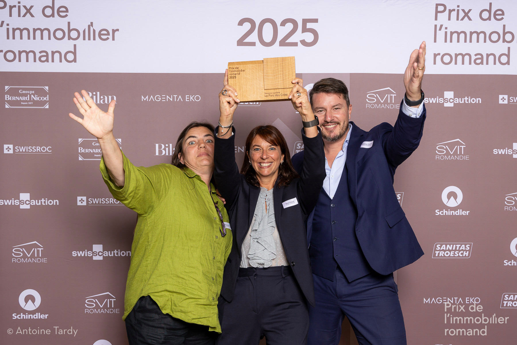 Trois personnes souriantes posent avec un trophée lors du Prix de l’immobilier romand 2025, devant un mur avec des logos d’entreprises.