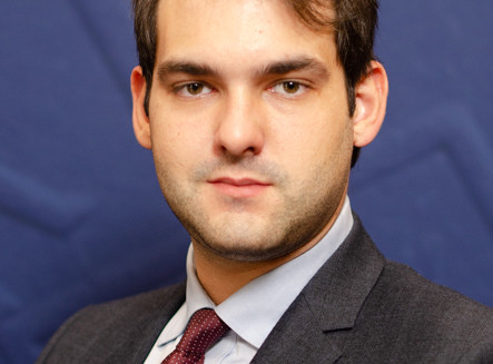 Jeune homme en costume gris et cravate bordeaux, photographié sur fond bleu foncé, portrait professionnel.