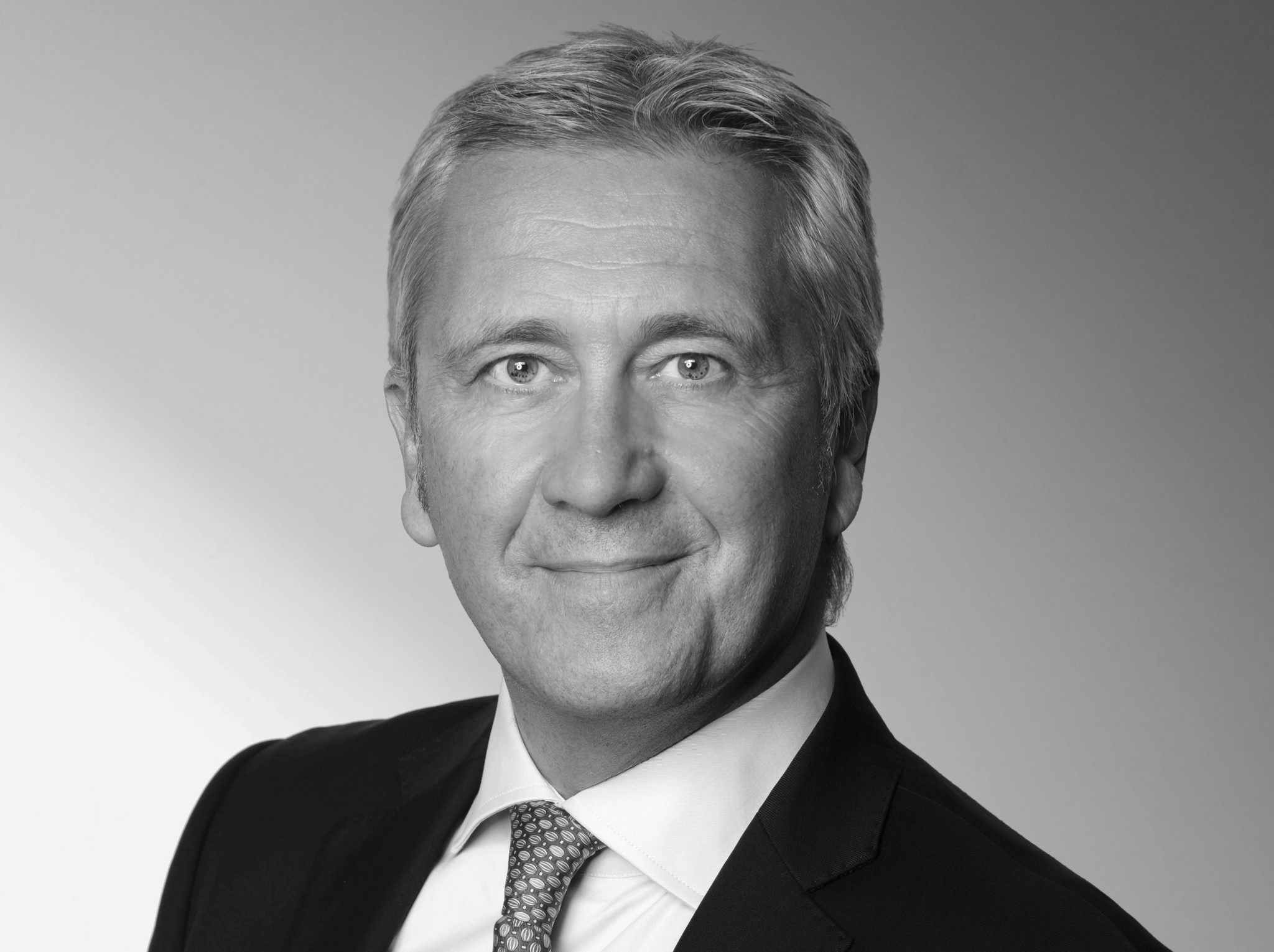 Portrait en noir et blanc d’un homme d’affaires en costume sombre et cravate, souriant face à l’objectif.