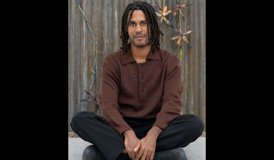 Un homme aux dreadlocks portant un pull marron, assis en tailleur devant un mur métallique avec des éléments colorés.