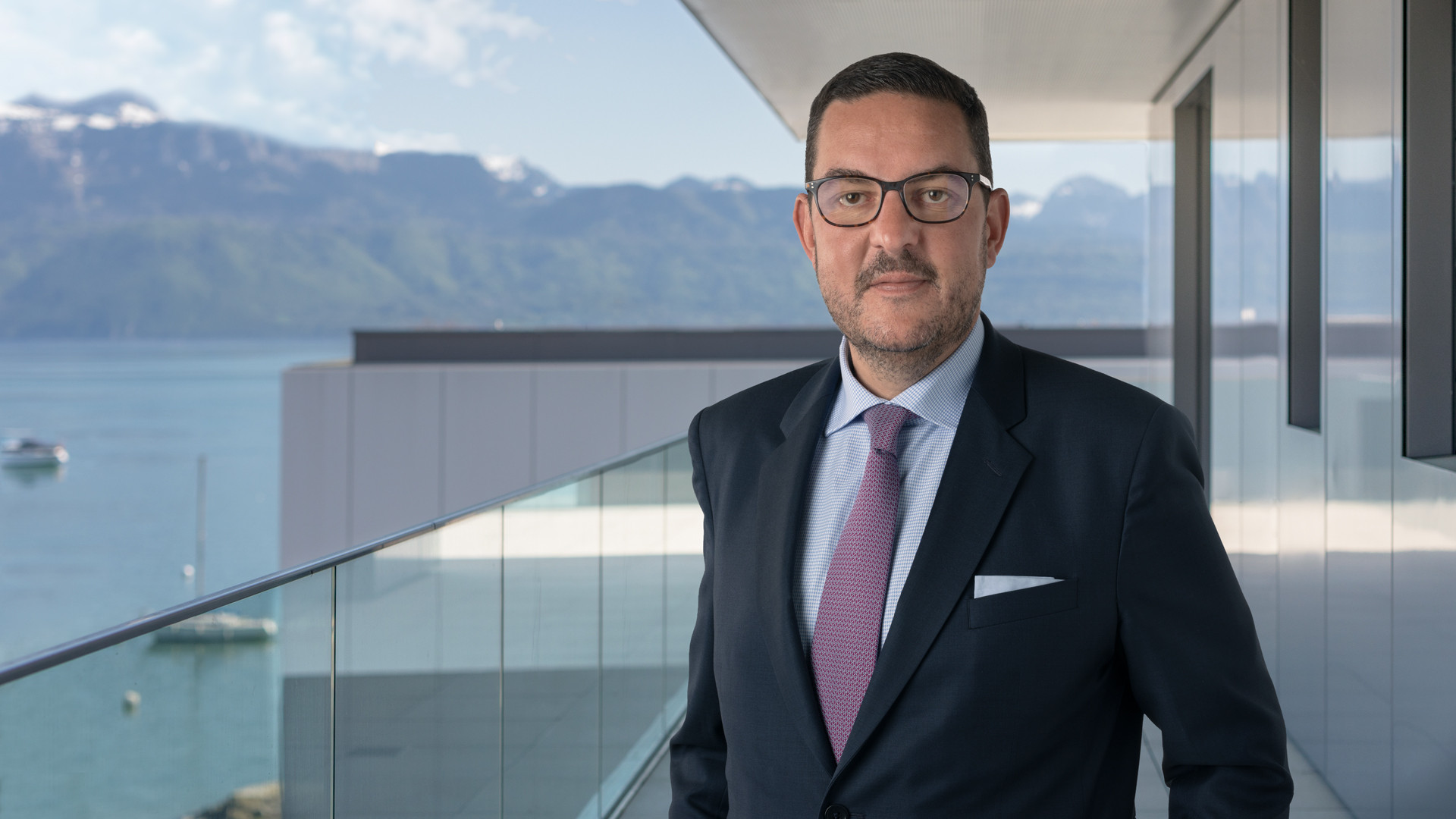 Homme en costume sombre et cravate rose posant sur un balcon avec vue sur un lac et des montagnes alpines