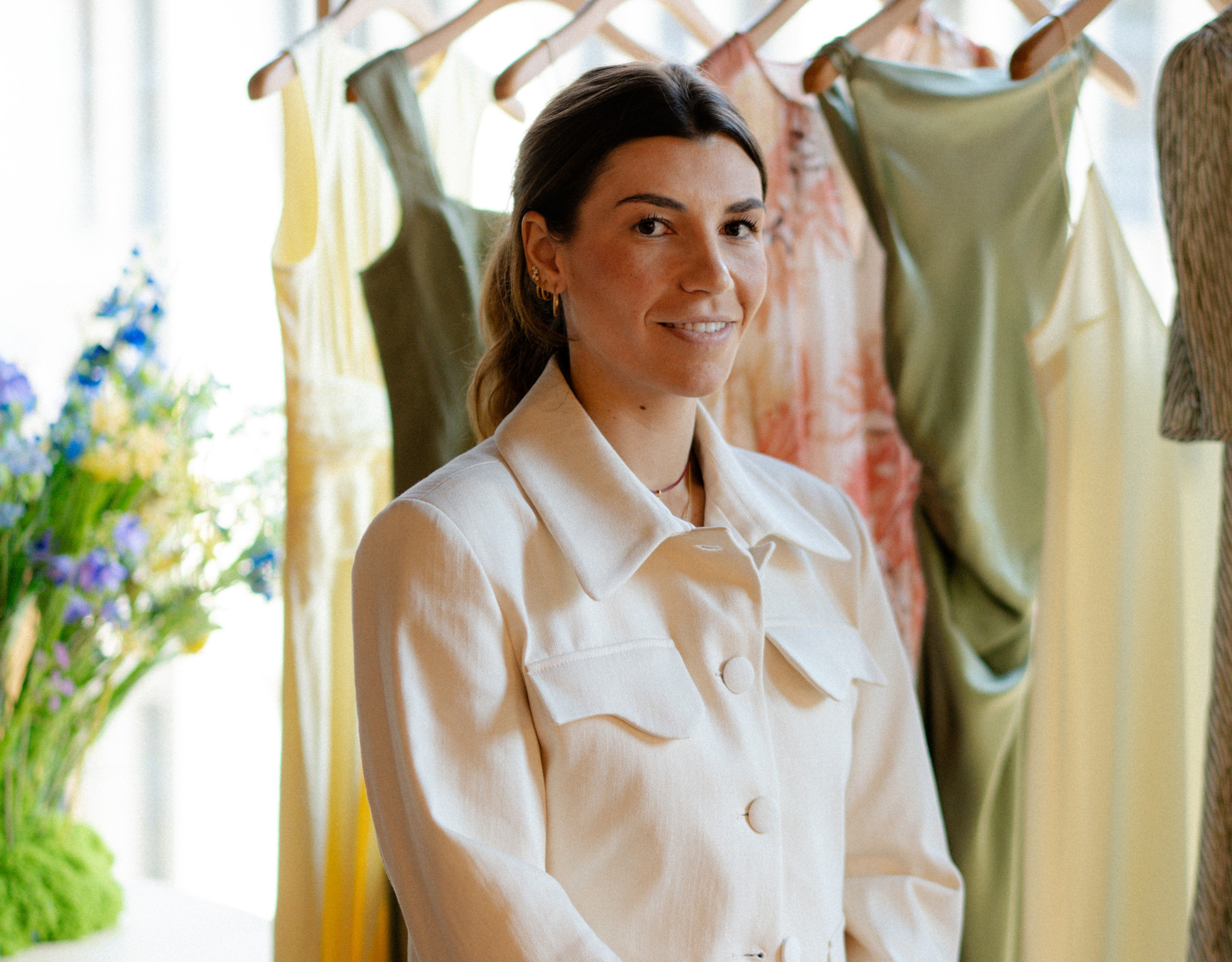 Jeune femme souriante en veste blanche devant un portant de robes colorées dans une boutique de mode.