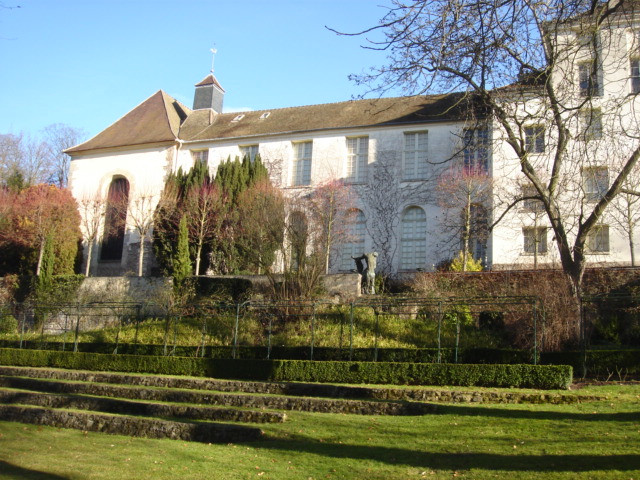 Bâtiment historique en pierre blanche entouré d’un jardin en terrasses avec haies taillées, arbres et ciel bleu hivernal.