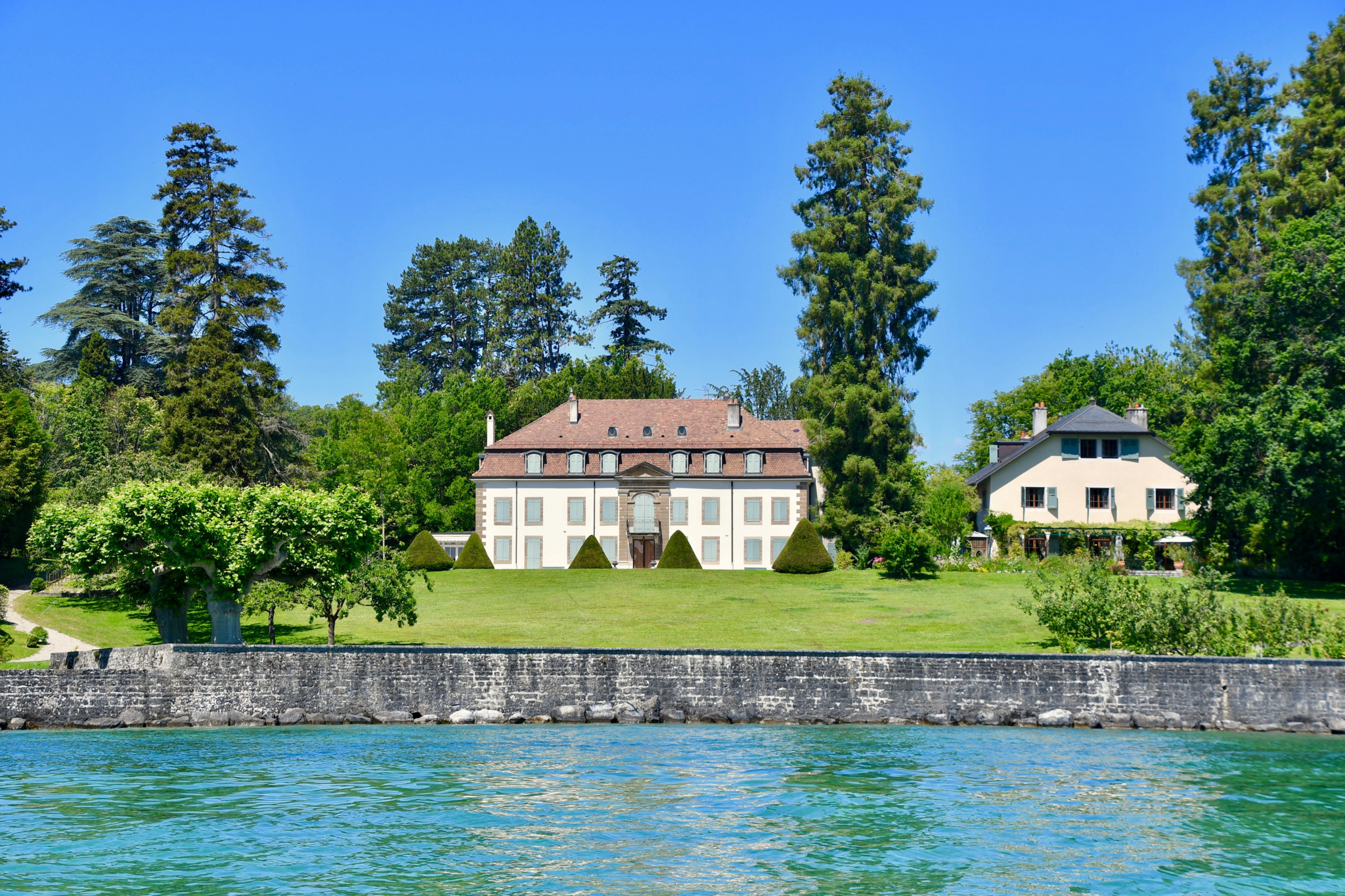 Élégante demeure historique au bord d’un lac aux eaux turquoise, entourée d’arbres et d’une pelouse verdoyante sous un ciel bleu.