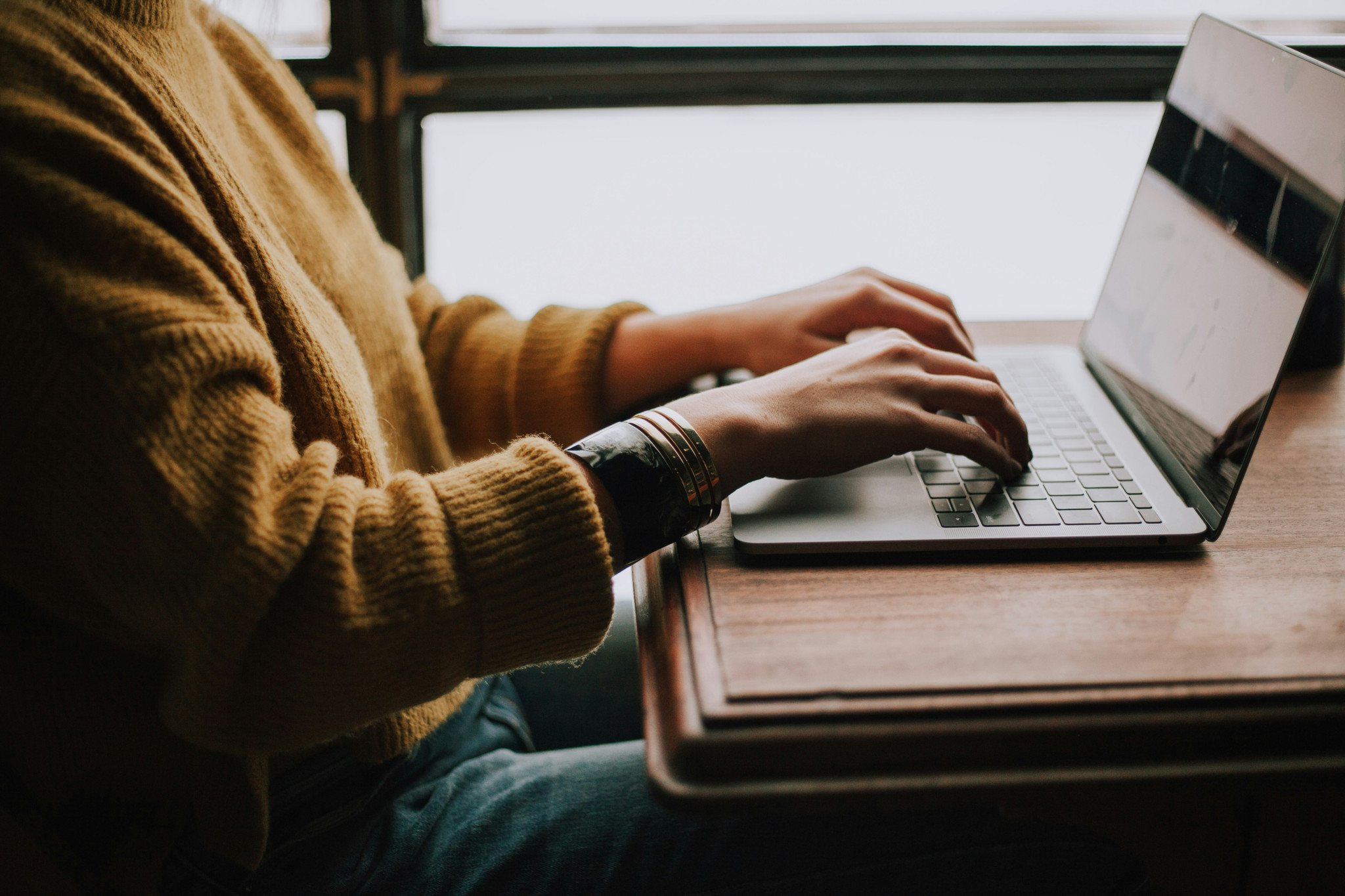 Personne en pull jaune moutarde tapant sur un ordinateur portable posé sur un bureau en bois près d’une fenêtre