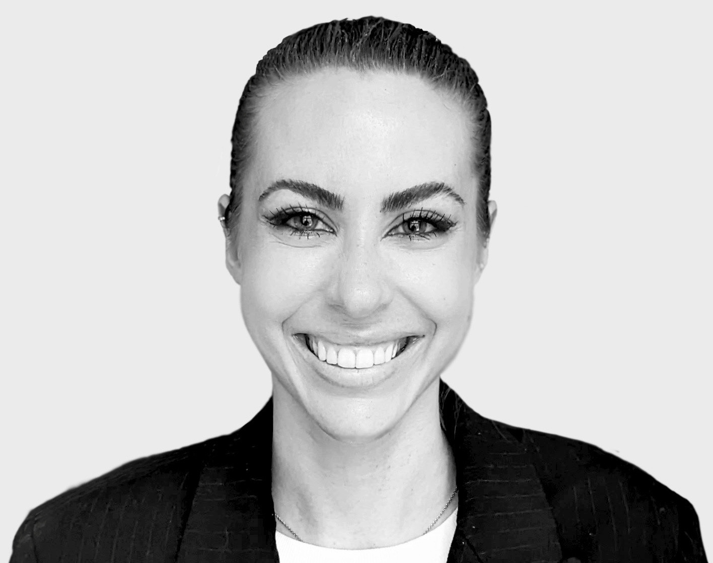 Portrait en noir et blanc d’une femme souriante aux cheveux tirés en arrière, portant un blazer sombre, sur fond gris clair.