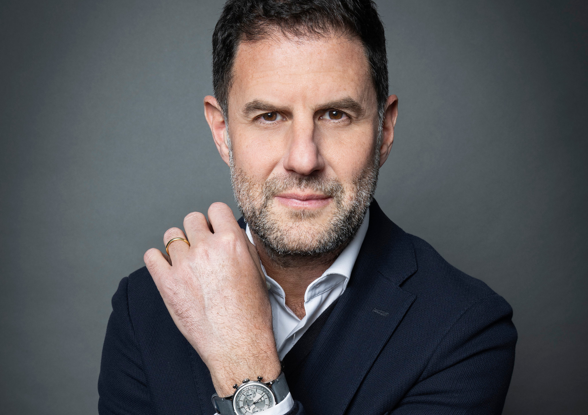 Homme en veston bleu marine et chemise blanche, portant une montre et une bague, posant devant un fond gris neutre.