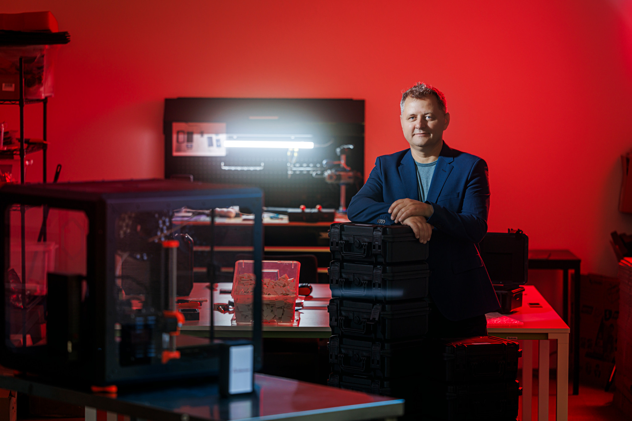 Un homme en veste bleue posant dans un atelier high-tech baigné de lumière rouge, entouré d’équipements électroniques et d’imprimantes 3D.