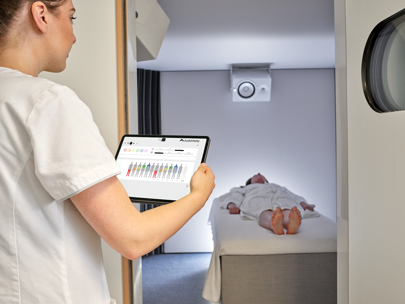 Une soignante consulte une tablette avec des données médicales colorées, tandis qu’un patient en peignoir repose sur un lit dans une chambre médicale.